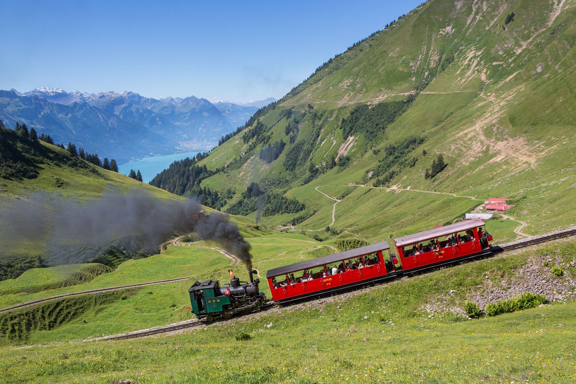 Steam train climbing a green mountain slope, with a lake and mountains in the distance.