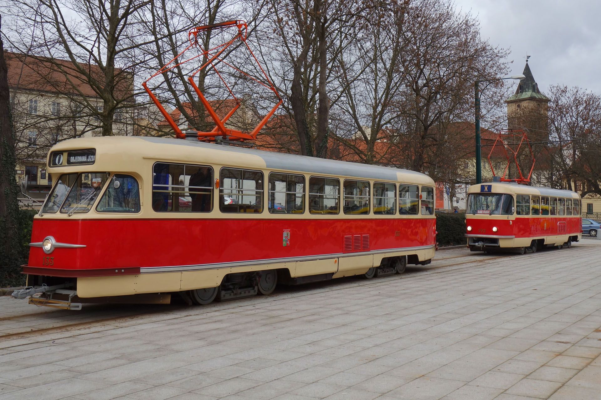 Red and cream-colored historic tram on tracks, another tram behind, in a town square.