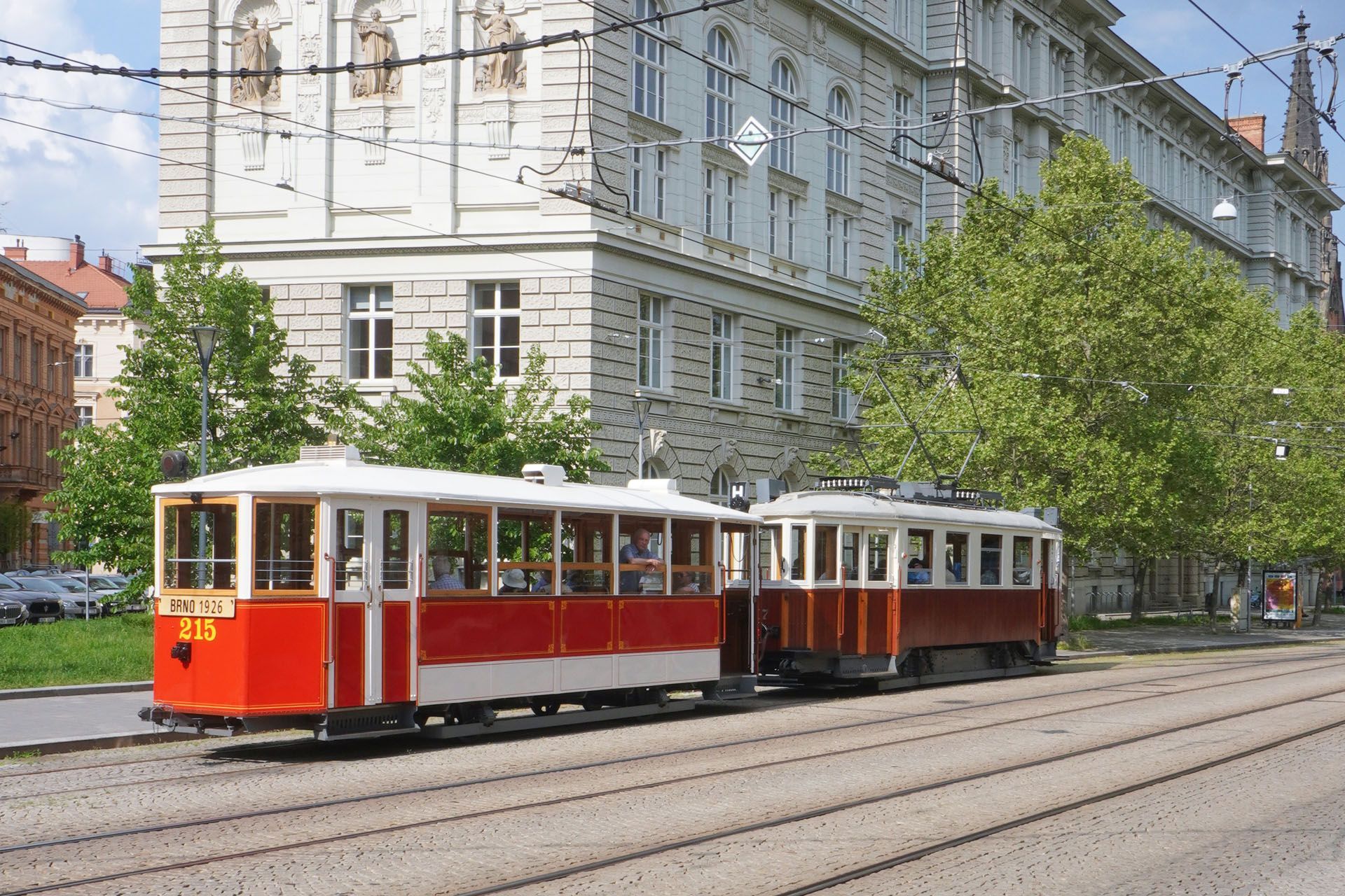 Two vintage red and white trams on tracks in a city, passing a large building.