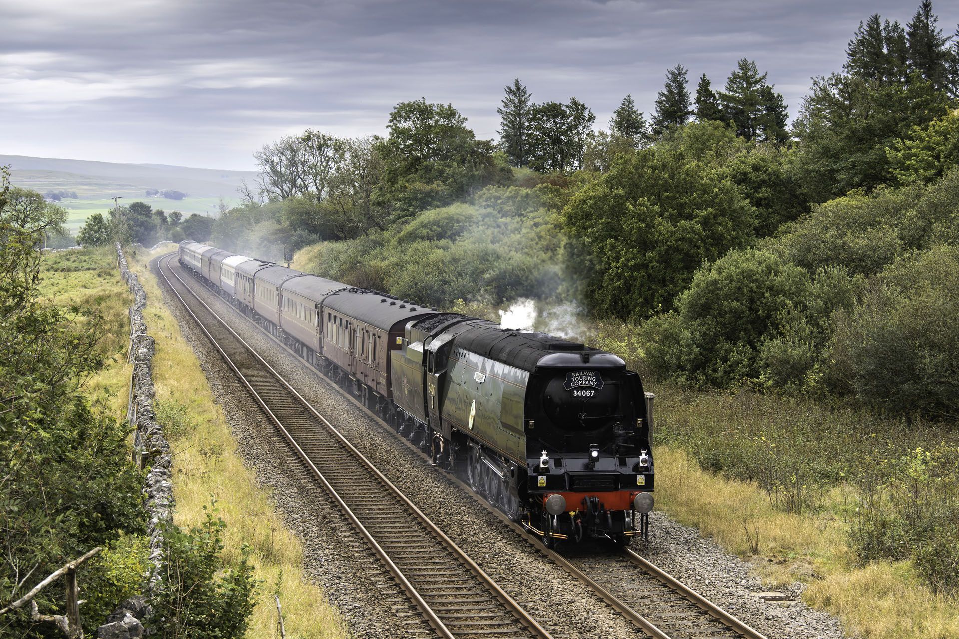 Steam train traveling on tracks through a green landscape, smoke rising from the engine.