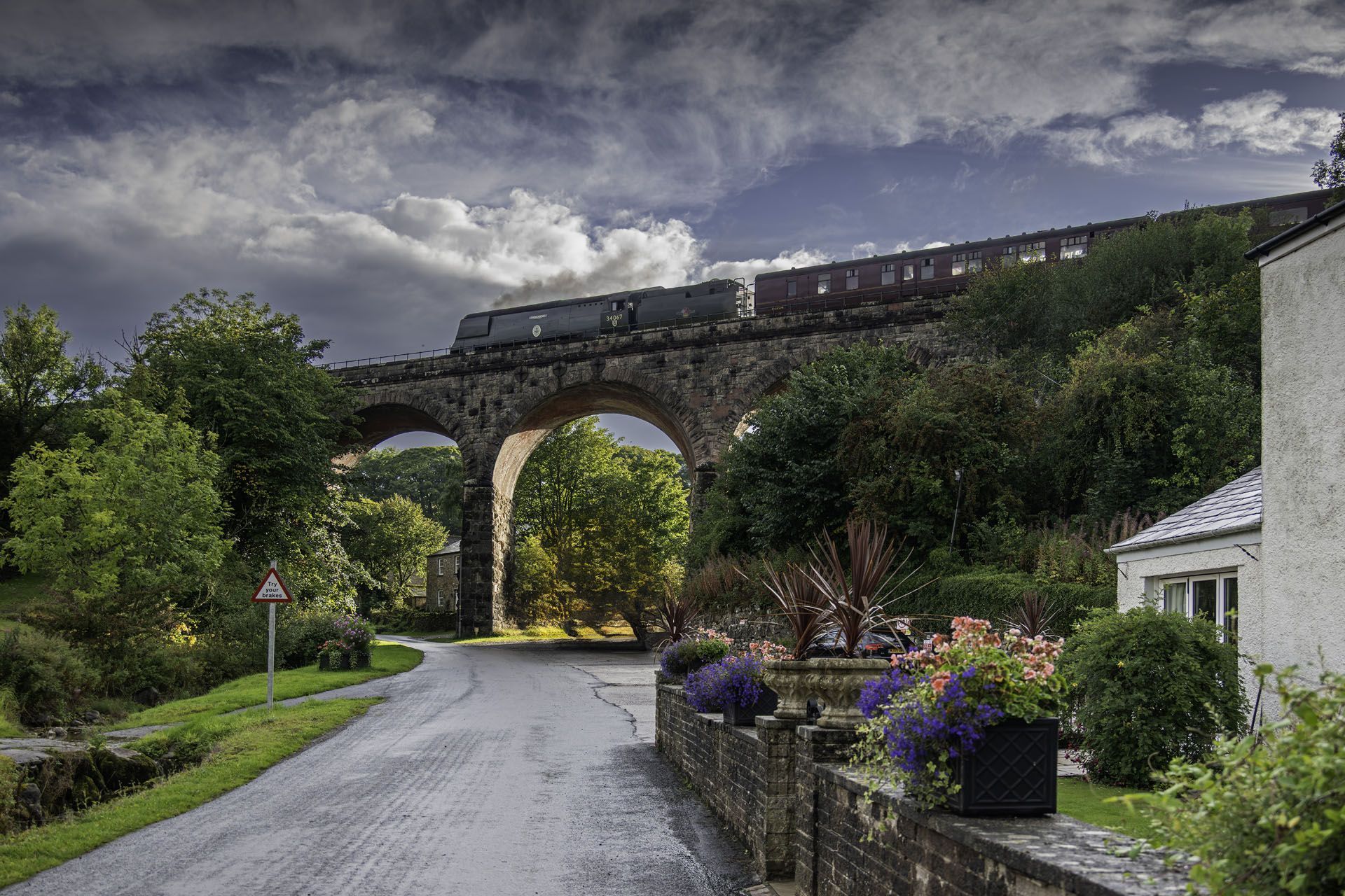 A train travels over a stone arch bridge on a cloudy day, road in the foreground, houses and trees on either side.