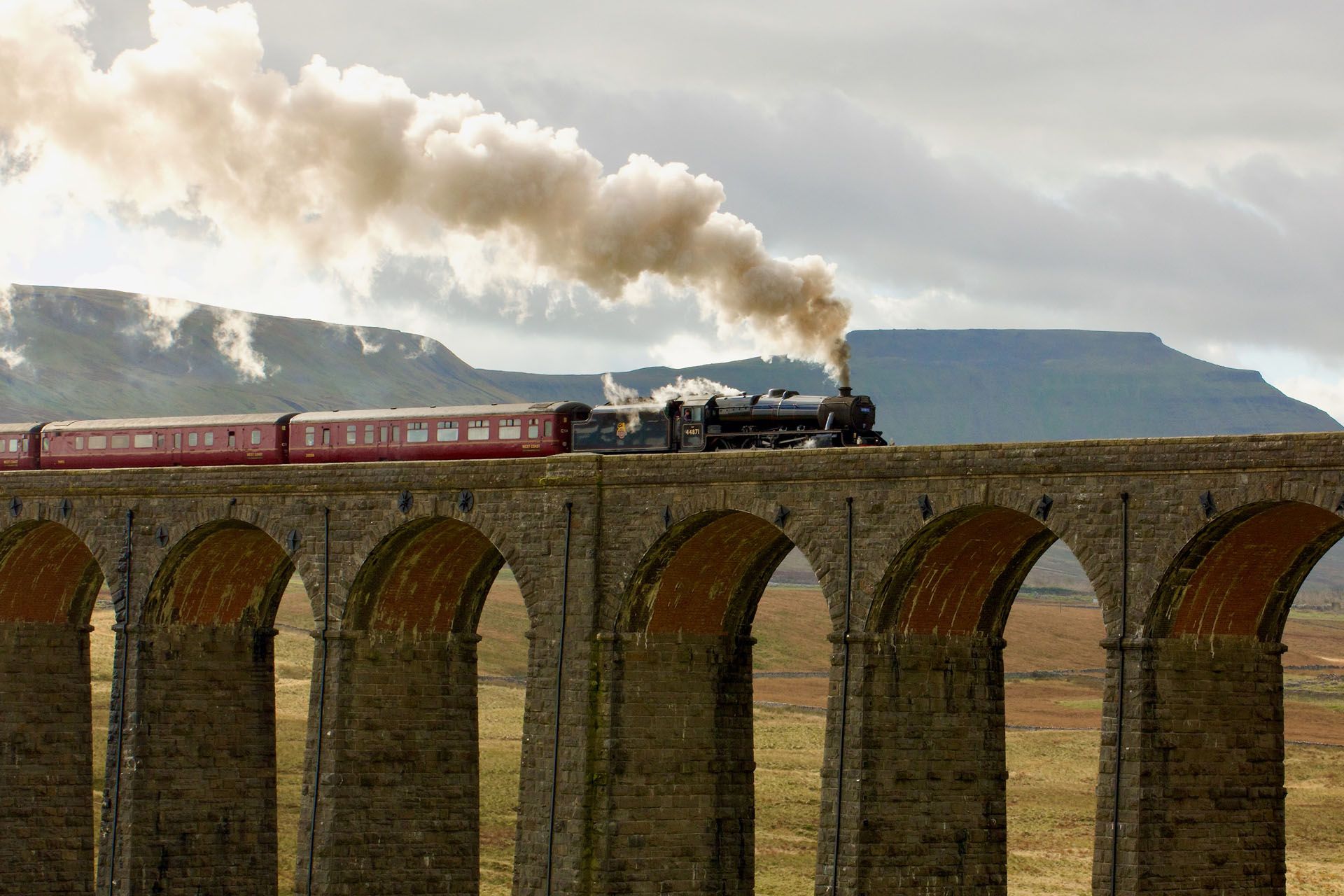 Steam train crossing a stone viaduct, smoke billowing into cloudy sky.