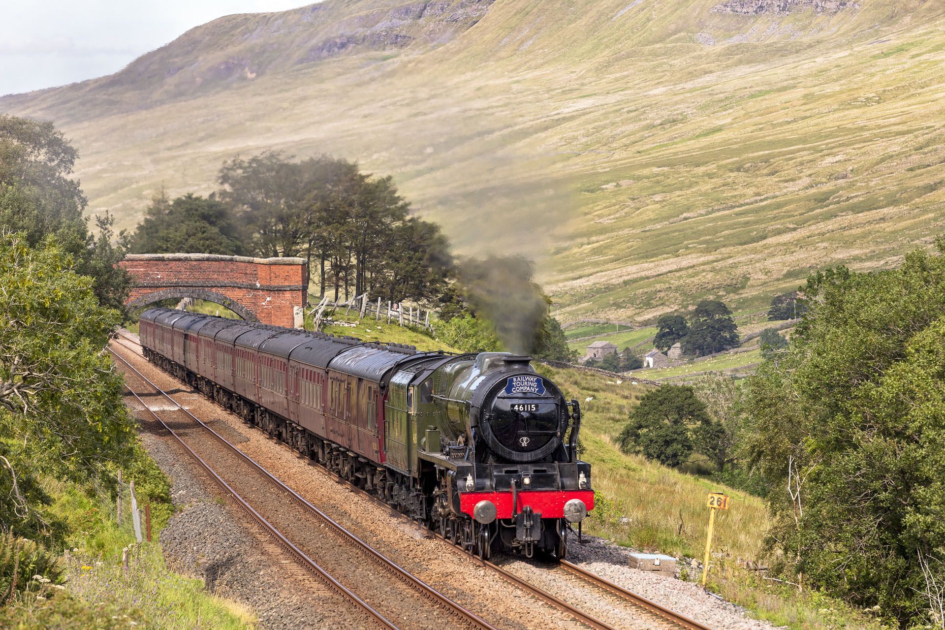 Steam train traveling on tracks through a green, hilly landscape.
