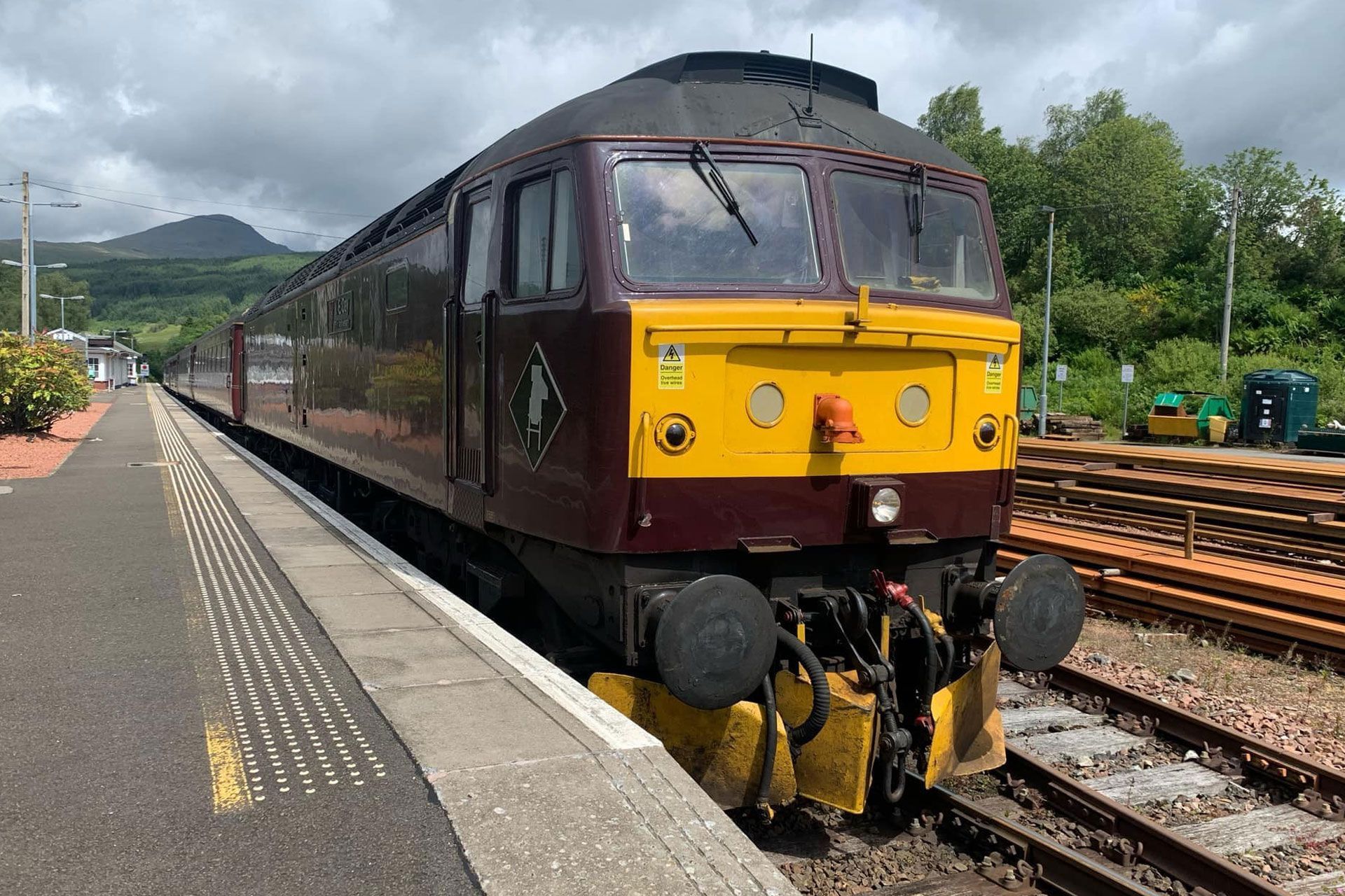 Maroon and yellow locomotive at a station platform with tracks and green hills in the background