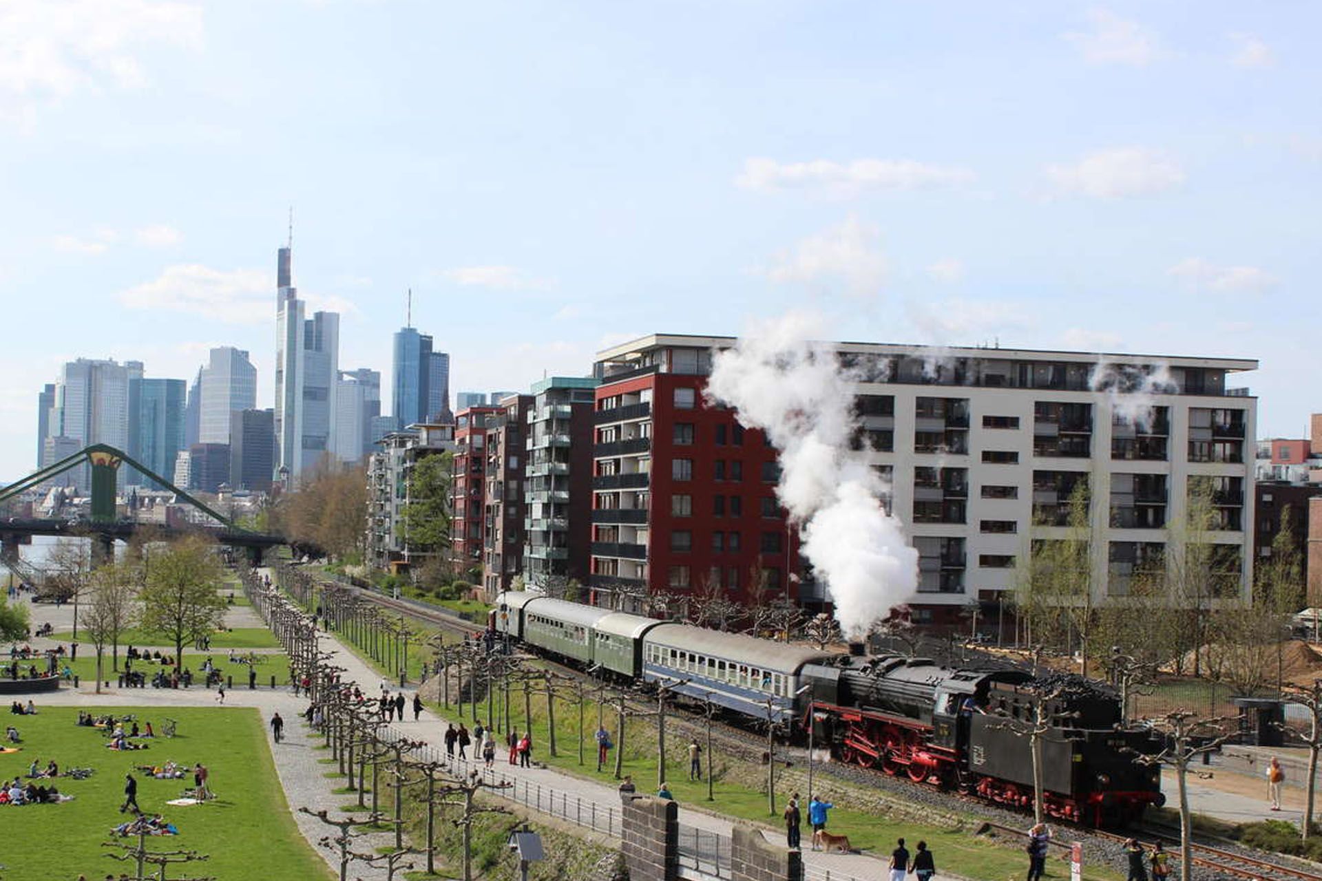 Steam locomotive pulling a train through a city park beside a skyline and river