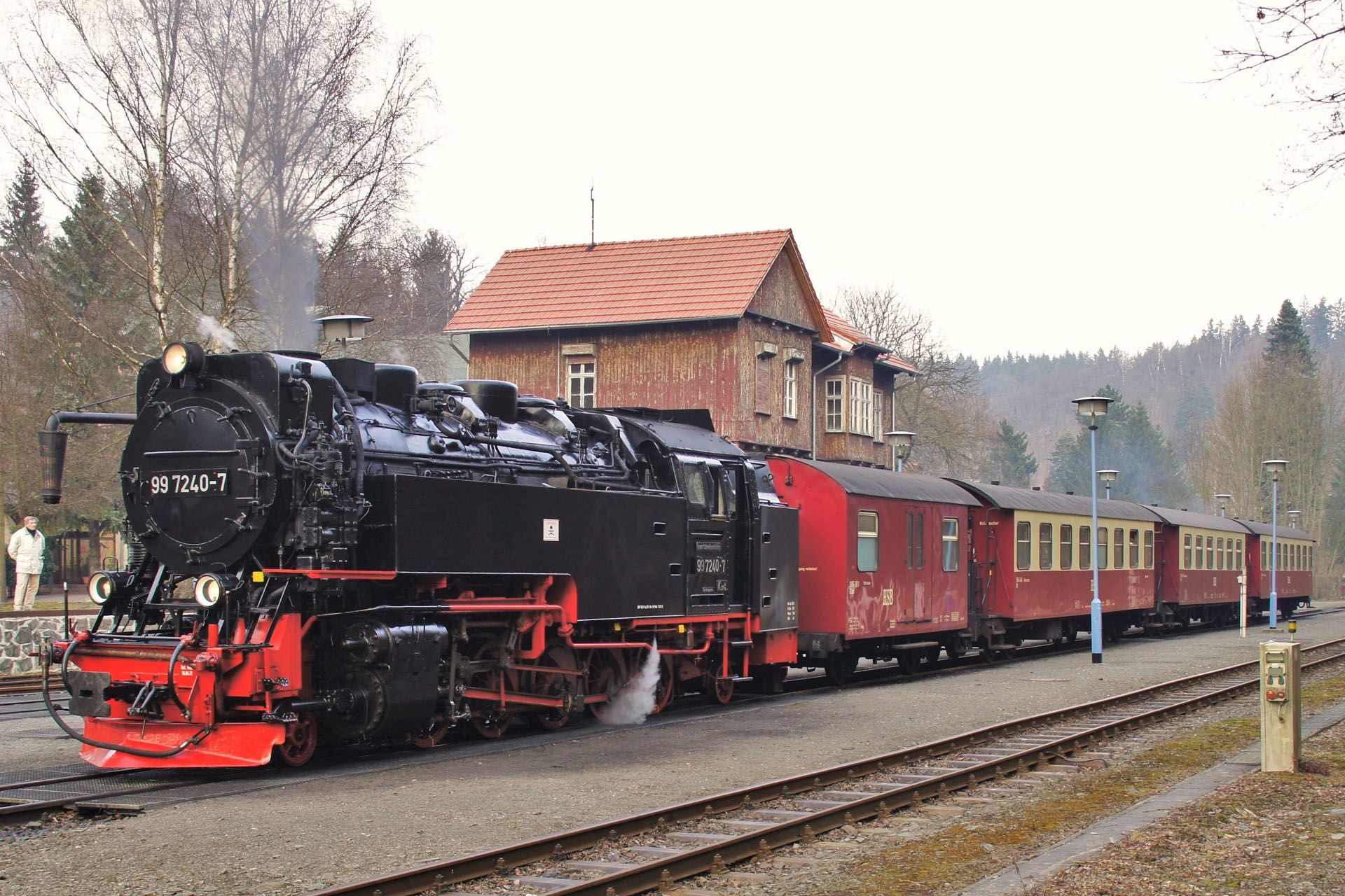 Black steam locomotive pulling red passenger cars beside a brick station in a wooded rural setting