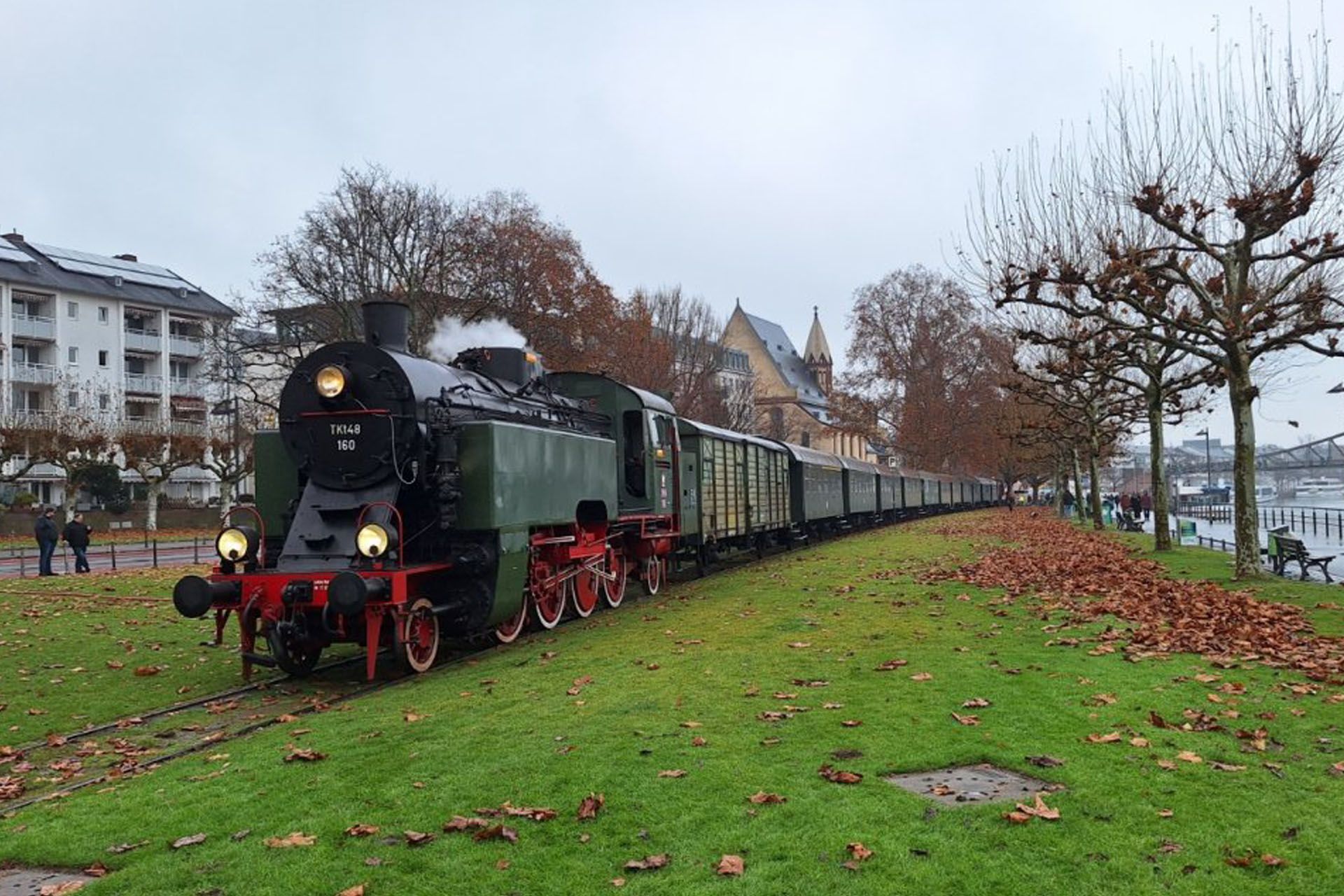Steam locomotive pulling passenger cars along a grassy riverside path in autumn