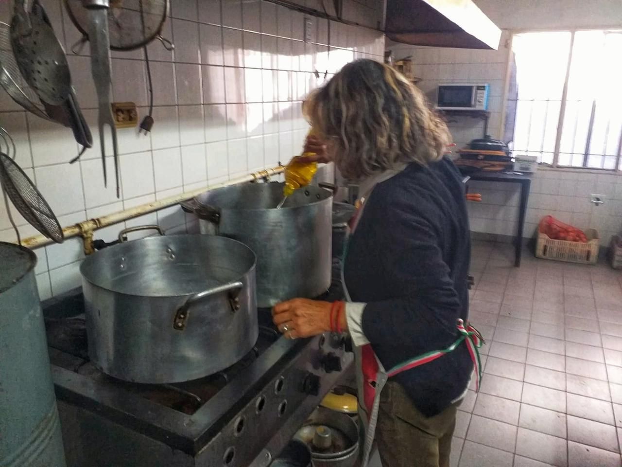 Una persona con delantal remueve una olla grande sobre la estufa en una cocina comercial con paredes alicatadas.