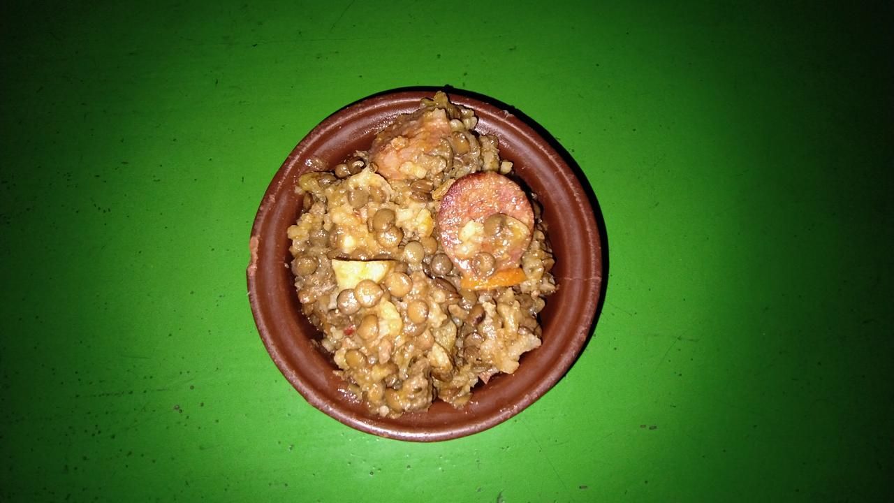 Un pequeño cuenco de arcilla marrón lleno de lentejas y rodajas de salchicha, centrado