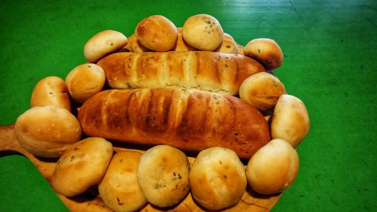 Dos hogazas de pan de color marrón dorado rodeadas de varios panecillos redondos sobre una tabla de madera, con fondo verde.