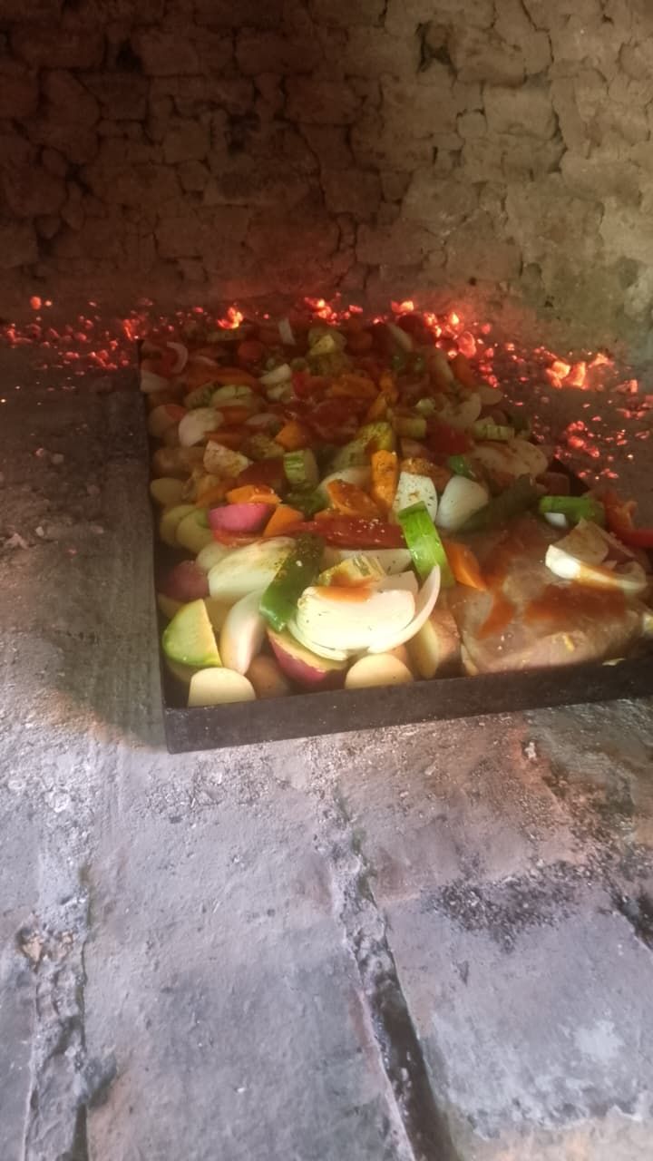 Una sartén metálica llena de verduras picadas y carne cocinándose dentro de un horno de piedra sobre brasas incandescentes.