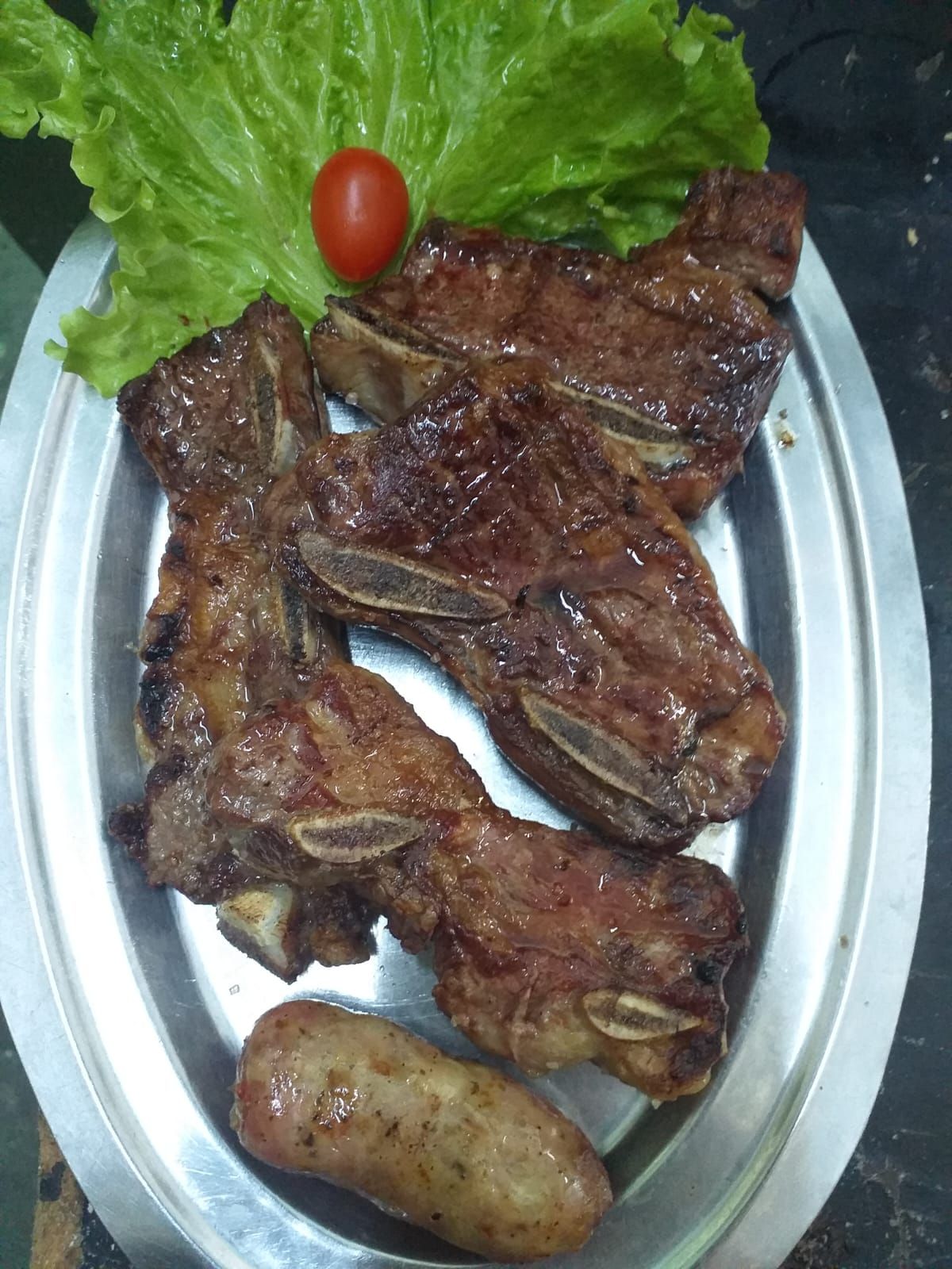 Una bandeja de plata contiene varios trozos de costillas de res a la parrilla, una salchicha, lechuga y un tomate cherry.