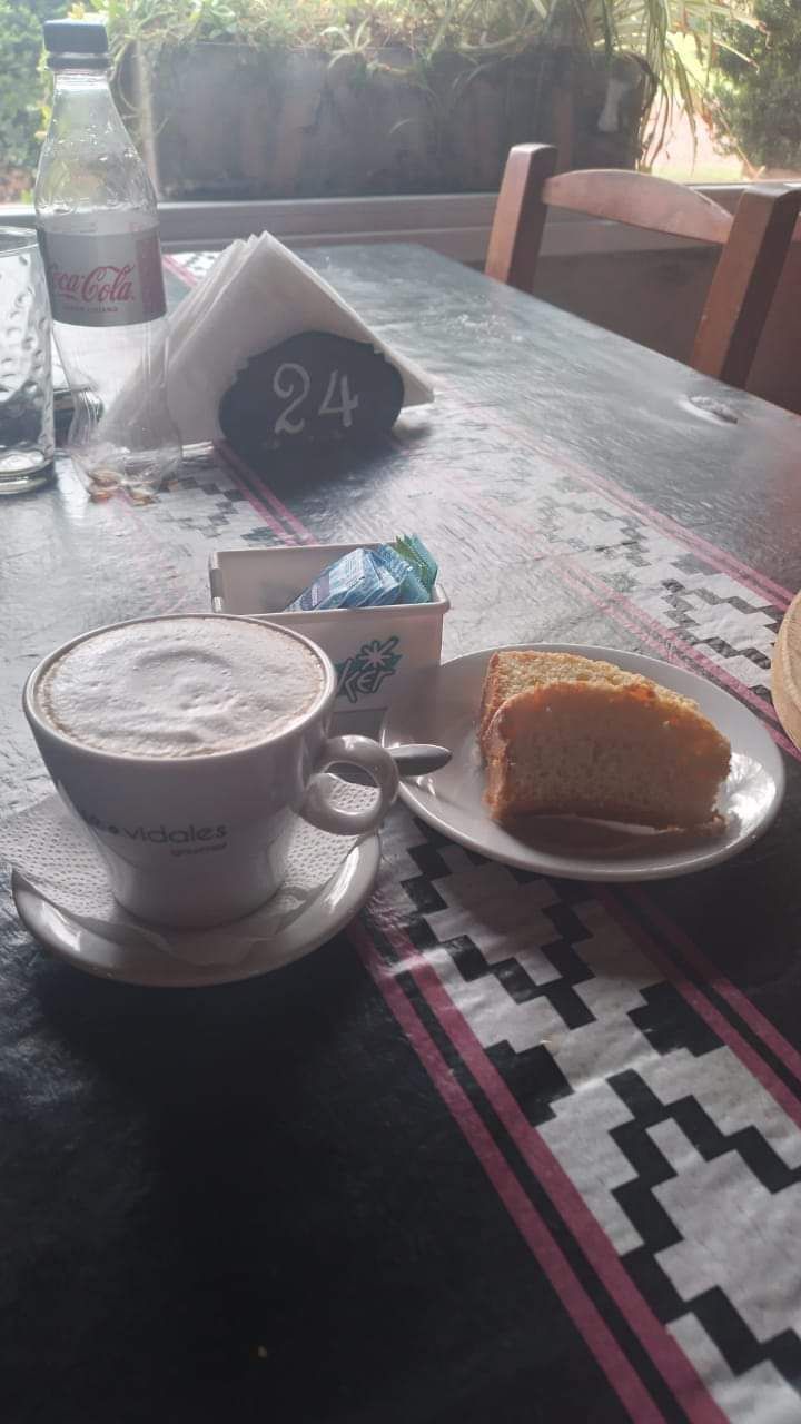 Un café con espuma en una taza blanca junto a una porción de pastel sobre un mantel 