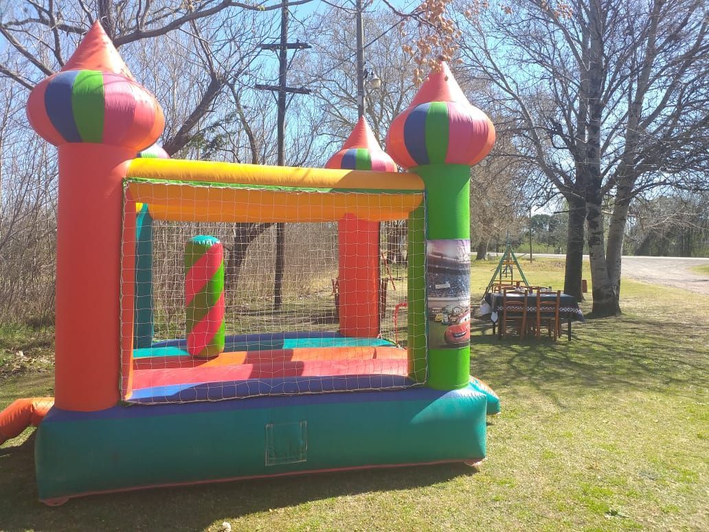 Un colorido castillo hinchable se alza sobre un campo de hierba con árboles y una mesa de picnic al fondo.