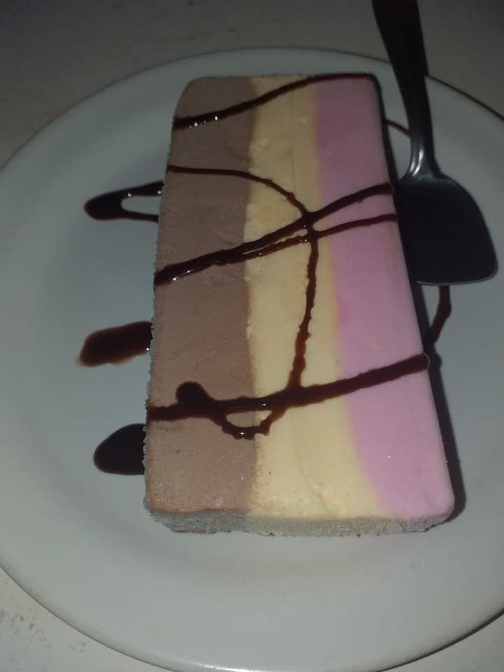 Una porción rectangular de helado tricolor —chocolate, vainilla y fresa— rociada con salsa de chocolate en un plato.