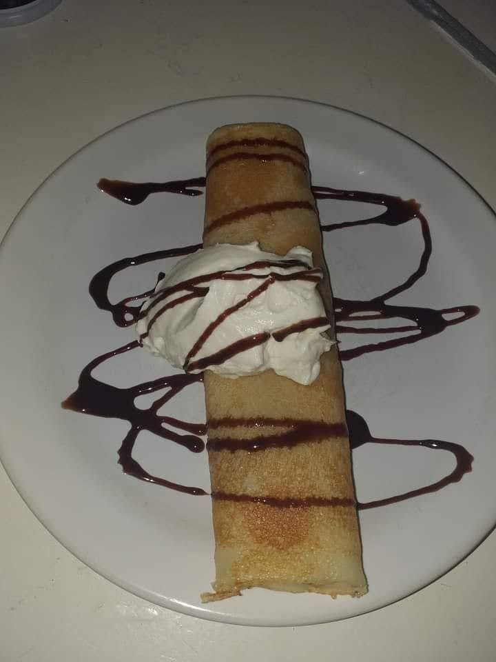 Una crêpe enrollada, cubierta con una cucharada de nata montada y rociada con salsa de chocolate, servida en un plato blanco.