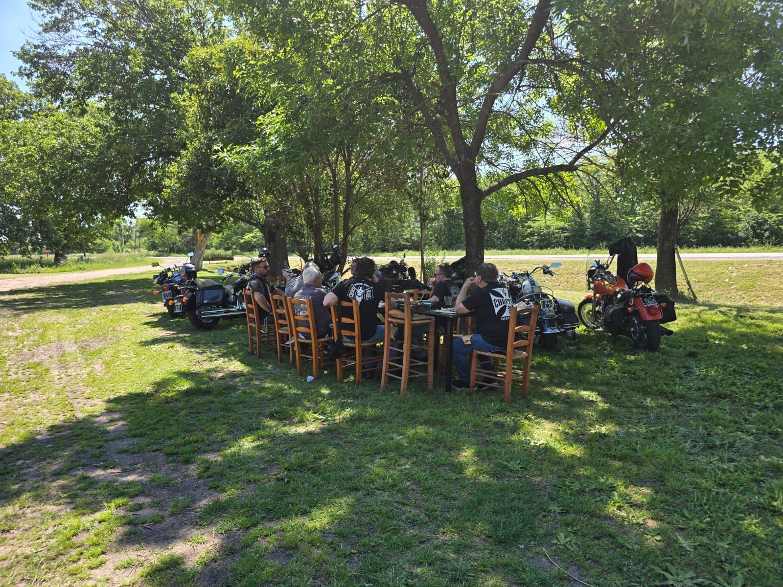 Un grupo de personas está sentado en una mesa de madera a la sombra de los árboles, con varias motocicletas