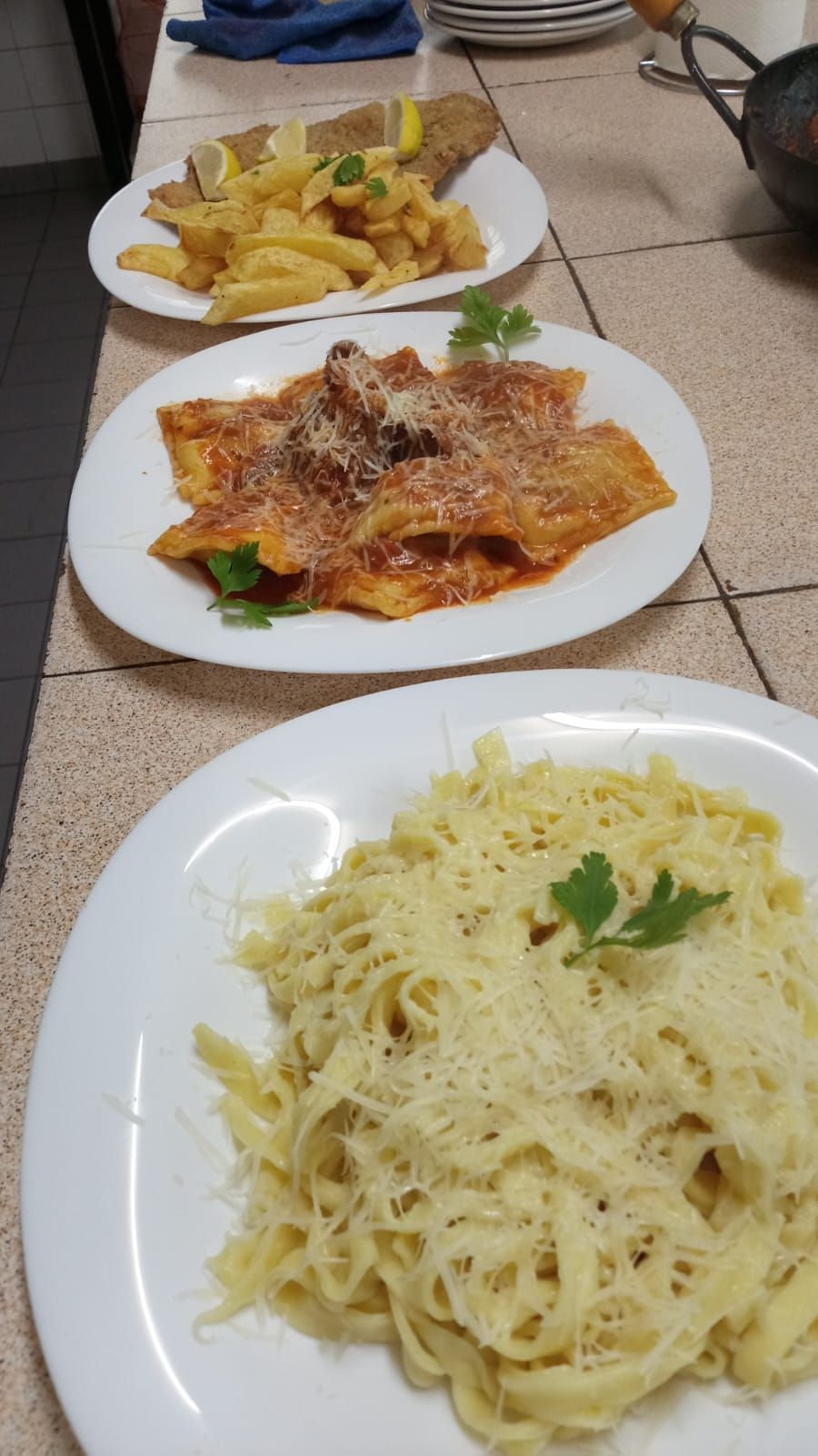 Tres platos de comida sobre un mostrador de azulejos: fettuccine Alfredo, pasta con salsa roja y pescado con patatas fritas.