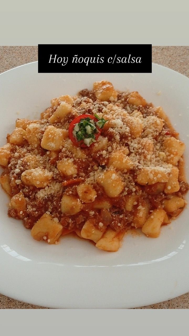 Un plato blanco lleno de ñoquis cubiertos con salsa de carne roja y espolvoreados con queso