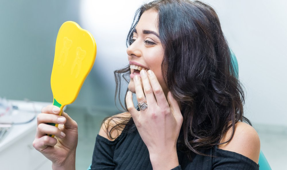 Une femme assise chez le dentiste examine ses dents dans un miroir à main jaune, souriant avec sa main près de sa bouche.