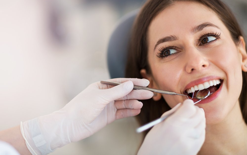 Une femme se fait examiner les dents chez le dentiste. Les mains gantées du dentiste tiennent des instruments dans sa bouche ; elle sourit.