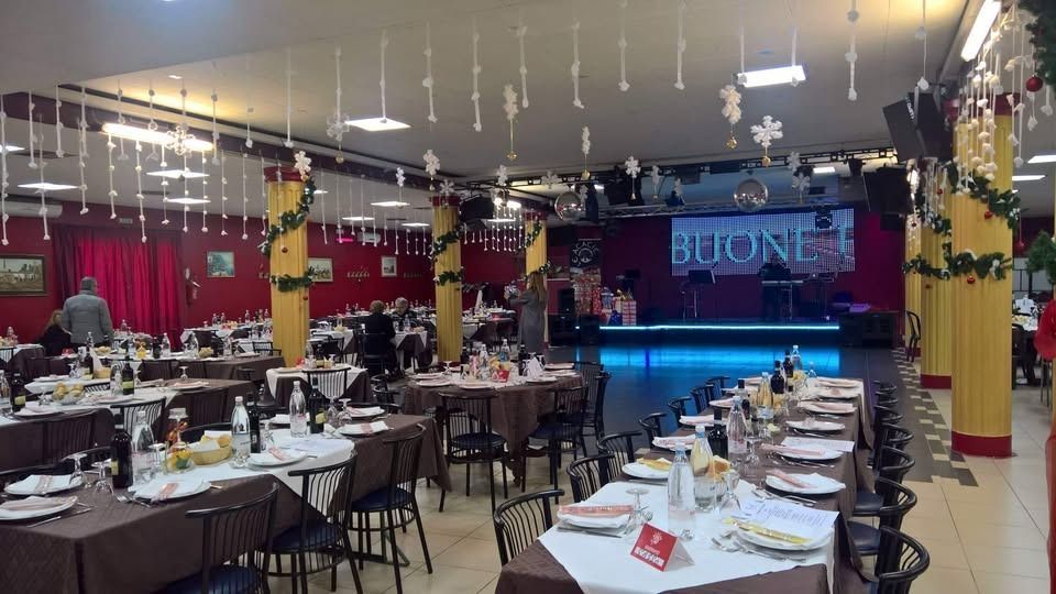 Sala da banchetto decorata con tavoli rotondi, tende rosse e un palco illuminato sullo sfondo.