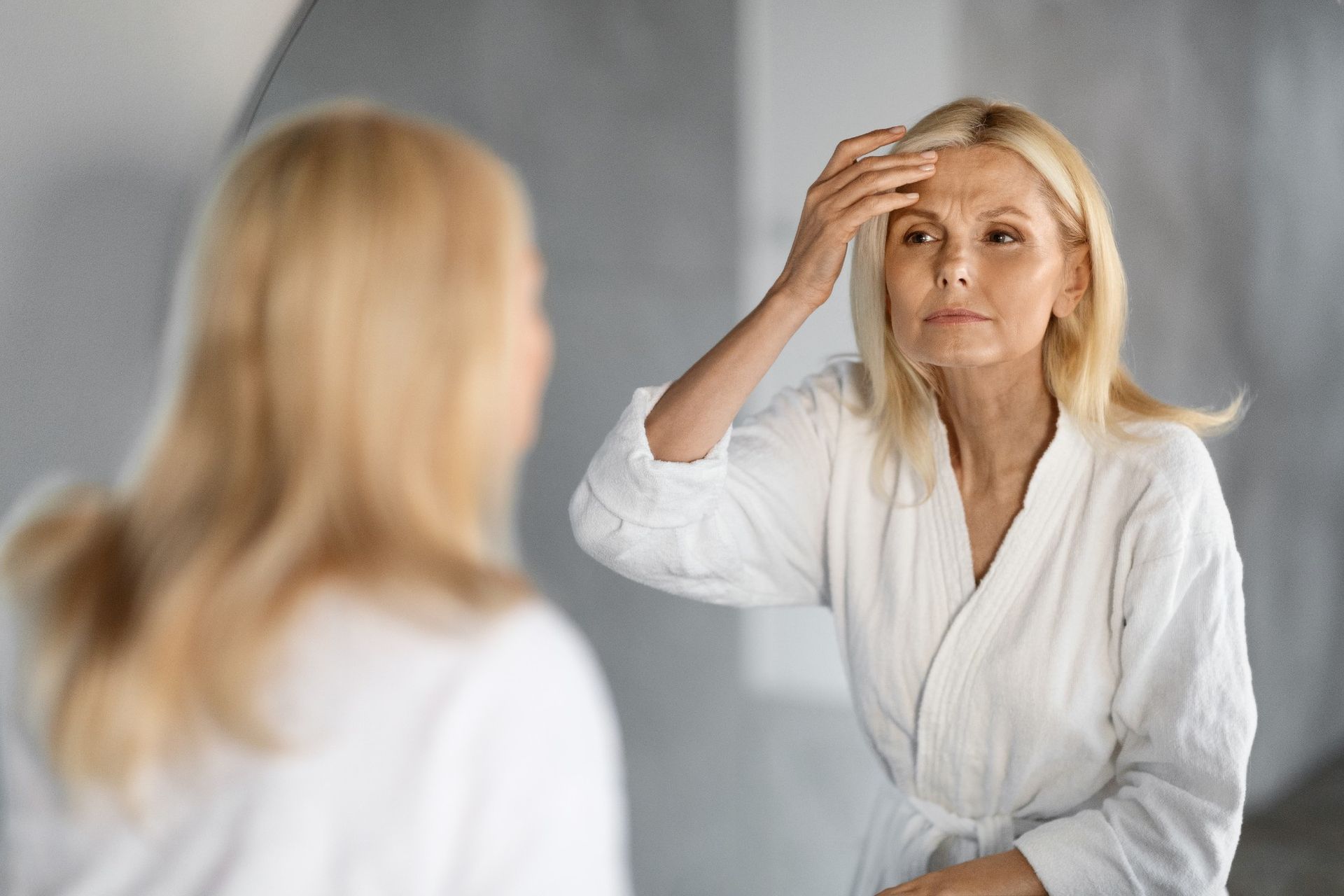 a woman in a bathrobe examines her forehead wrinkles in mirror