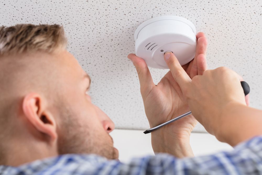 Man Installing A Smoke Detector On A White, Textured Ceiling With A Screwdriver — Stanayr Electrical in Gulliver, QLD