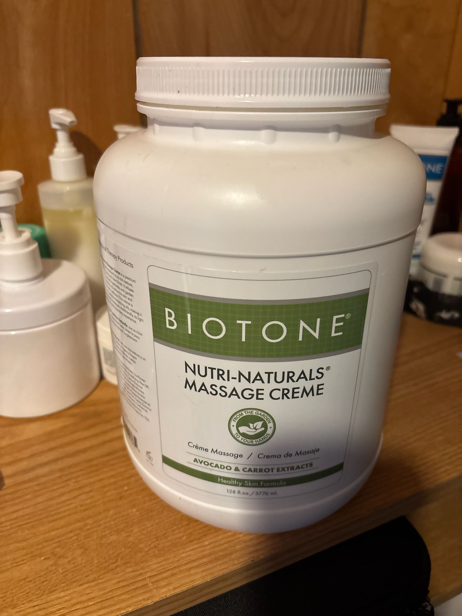 A large white tub of Biotone Nutri-Naturals Massage Creme sits on a wooden surface with other bottles in the background.