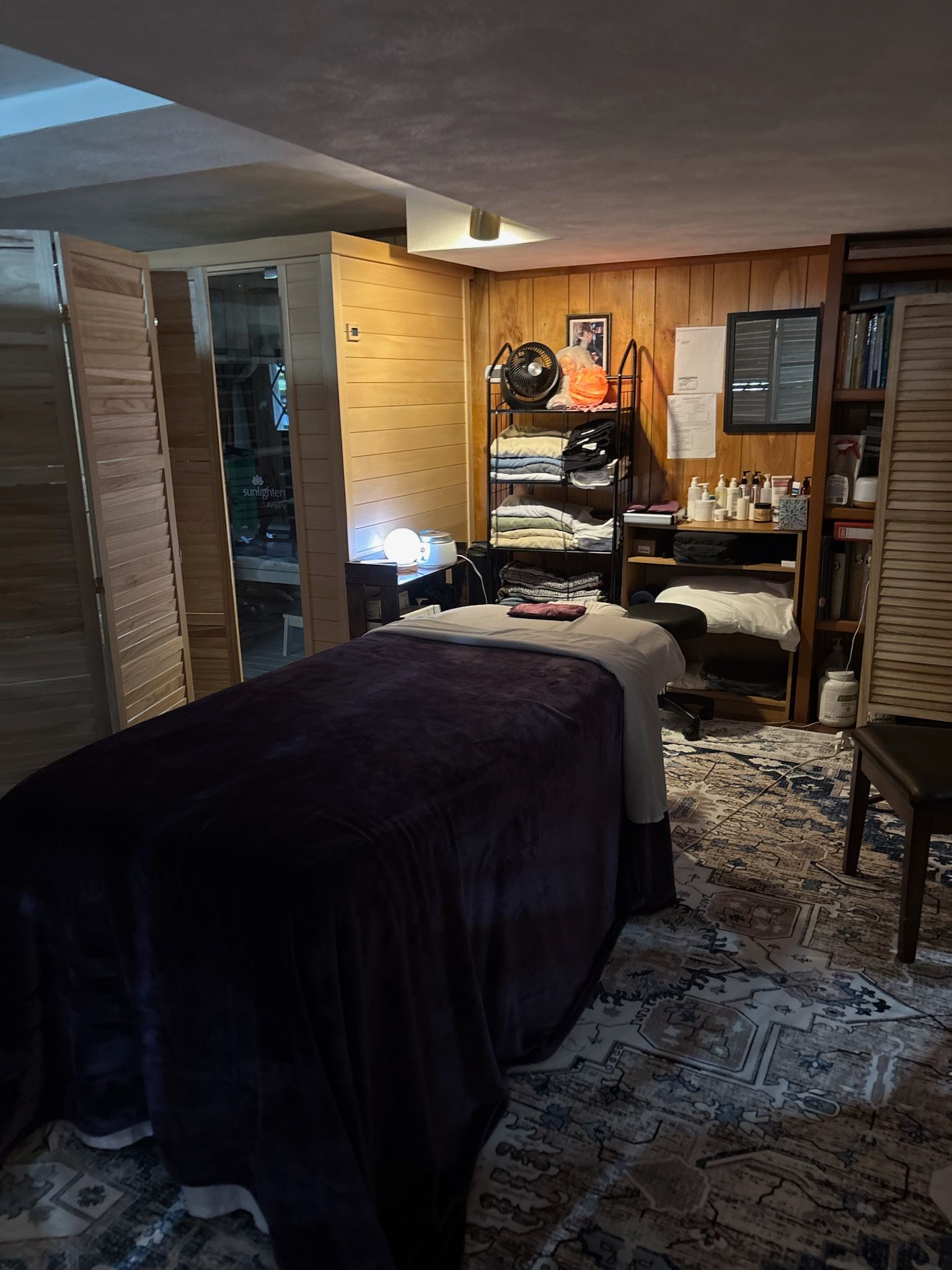 A spa treatment room featuring a massage table with a dark purple cover, wood paneled walls, and a bookshelf.