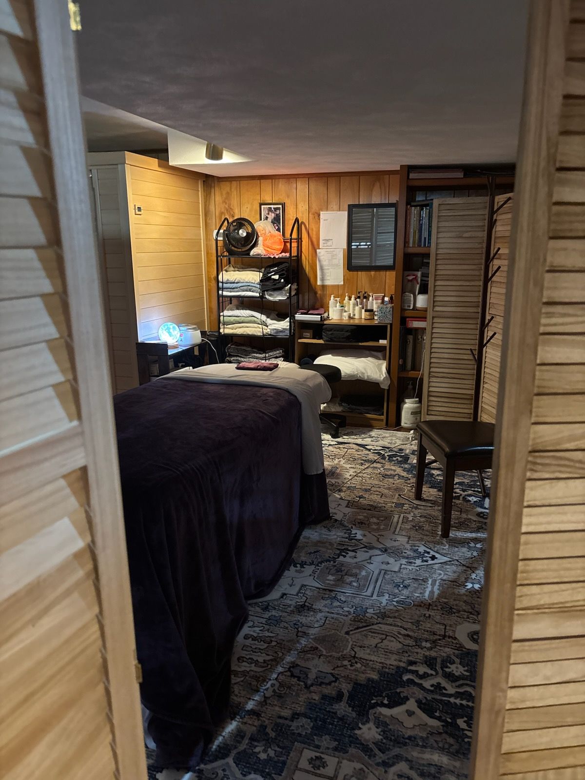 A dimly lit massage room featuring a covered table, a shelving unit with supplies, and a patterned rug on the floor.