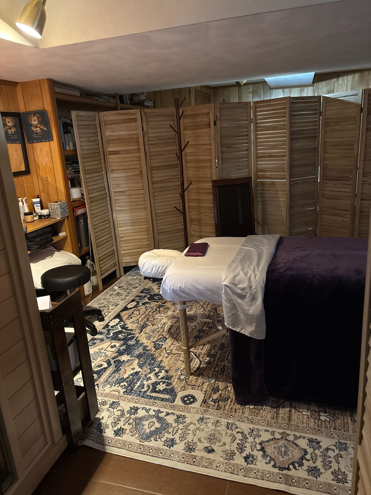 A massage therapy room with a treatment table covered in a white sheet and purple blanket, on a patterned blue-grey rug.