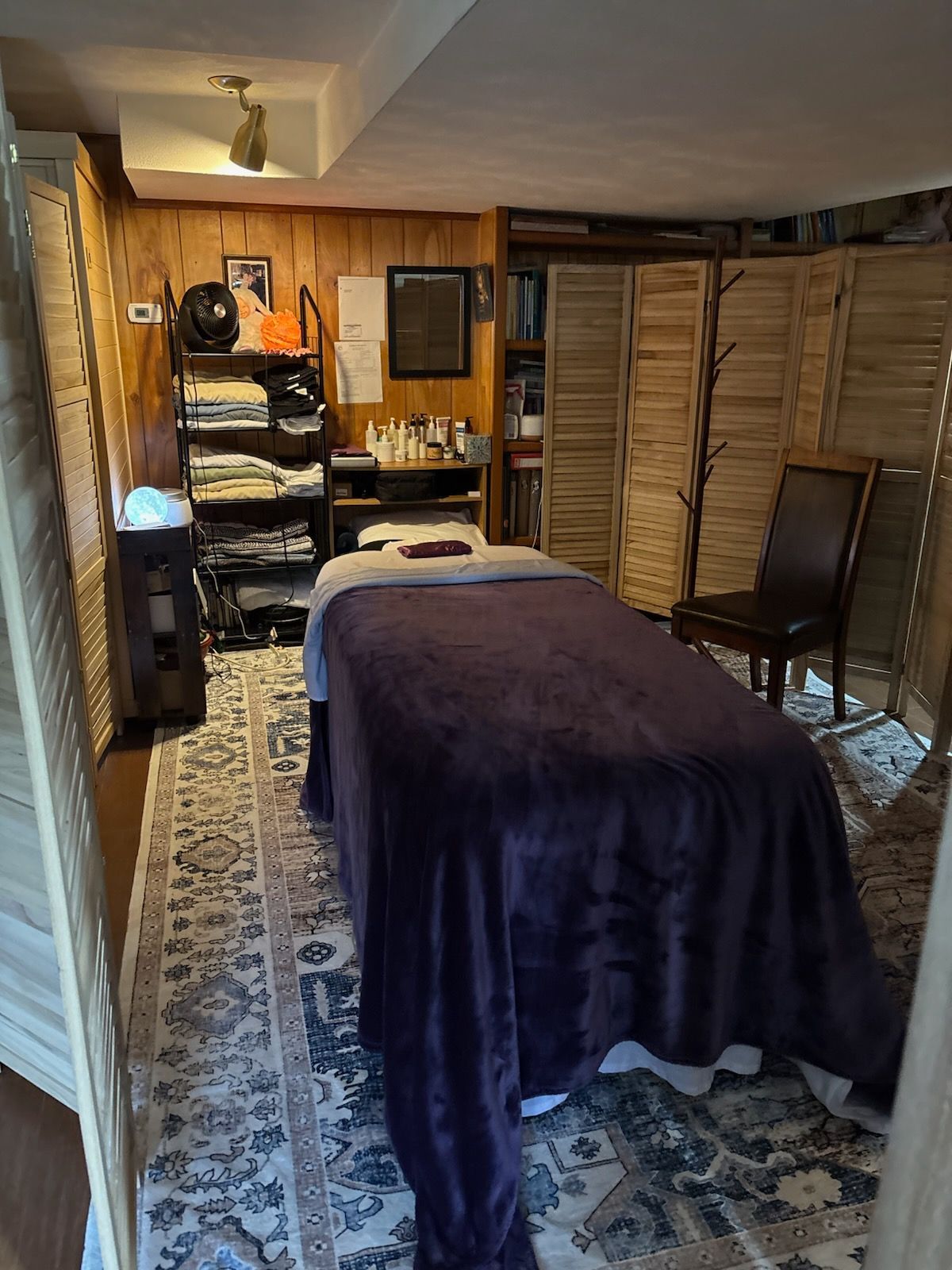 A massage therapy room with a treatment table covered in a dark purple blanket, a wooden chair, and a shelving unit.