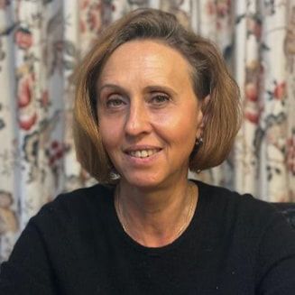 A person with chin-length hair wearing a black shirt, posing in front of a patterned curtain.