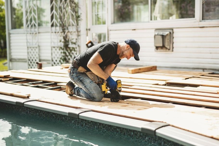 Worker kneeling on a pool deck, cutting wooden planks with a power tool beside the water.