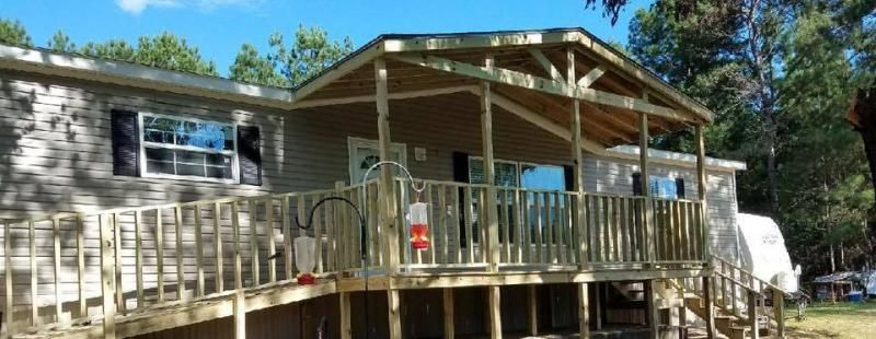 A mobile home with a large, elevated wooden porch and railing, set against a backdrop of trees under a blue sky.