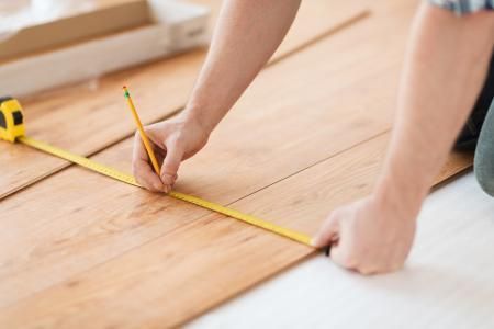 A person marks a measurement on light-colored wood floor planks with a yellow pencil and a tape measure.
