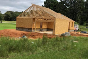 A house under construction with exposed wooden roof trusses and plywood walls, set on a rural site with red soil.