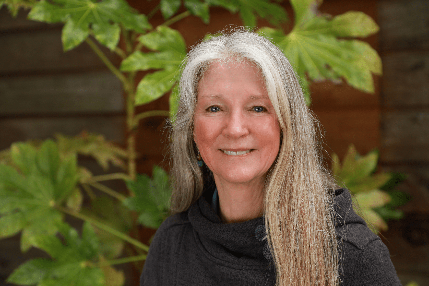 Woman with long gray hair smiles in front of green leaves and a wooden background.