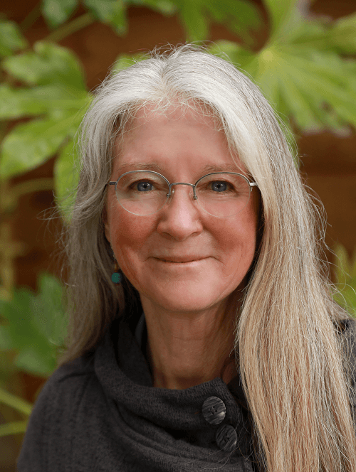A woman with long gray hair and glasses is smiling for the camera.