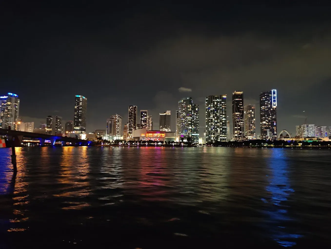 Night view of the Downtown Miami skyline from Biscayne Bay featuring Kaseya Center, the Signature Bridge, illuminated skyscrapers, and colorful waterfront reflections.