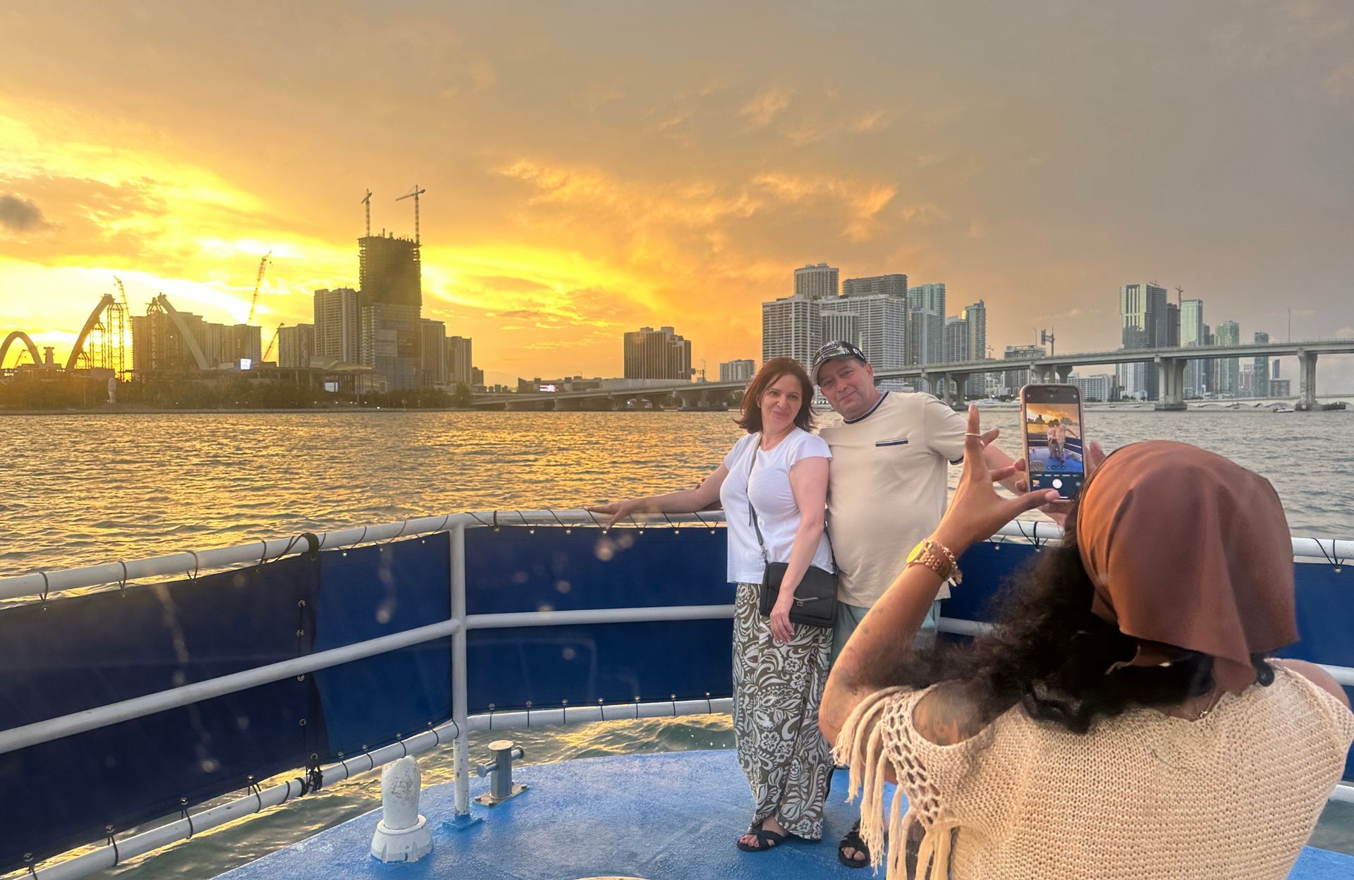 Couple posing for a photo on the front of the boat during a Miami Sightseeing Sunset Cruise with the downtown Miami skyline glowing at sunset over Biscayne Bay.
