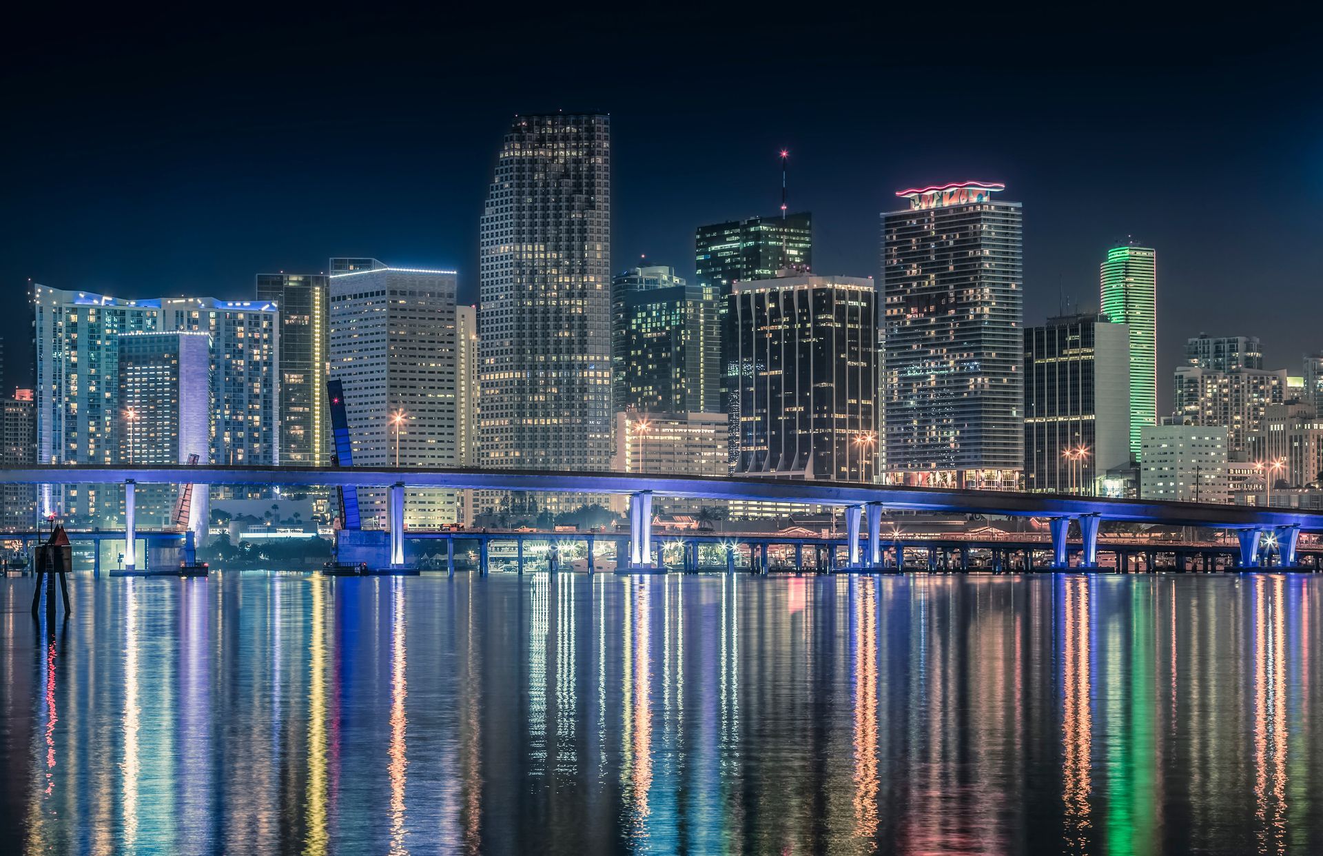 Miami skyline at night with downtown buildings and bridge lights reflecting on the water.