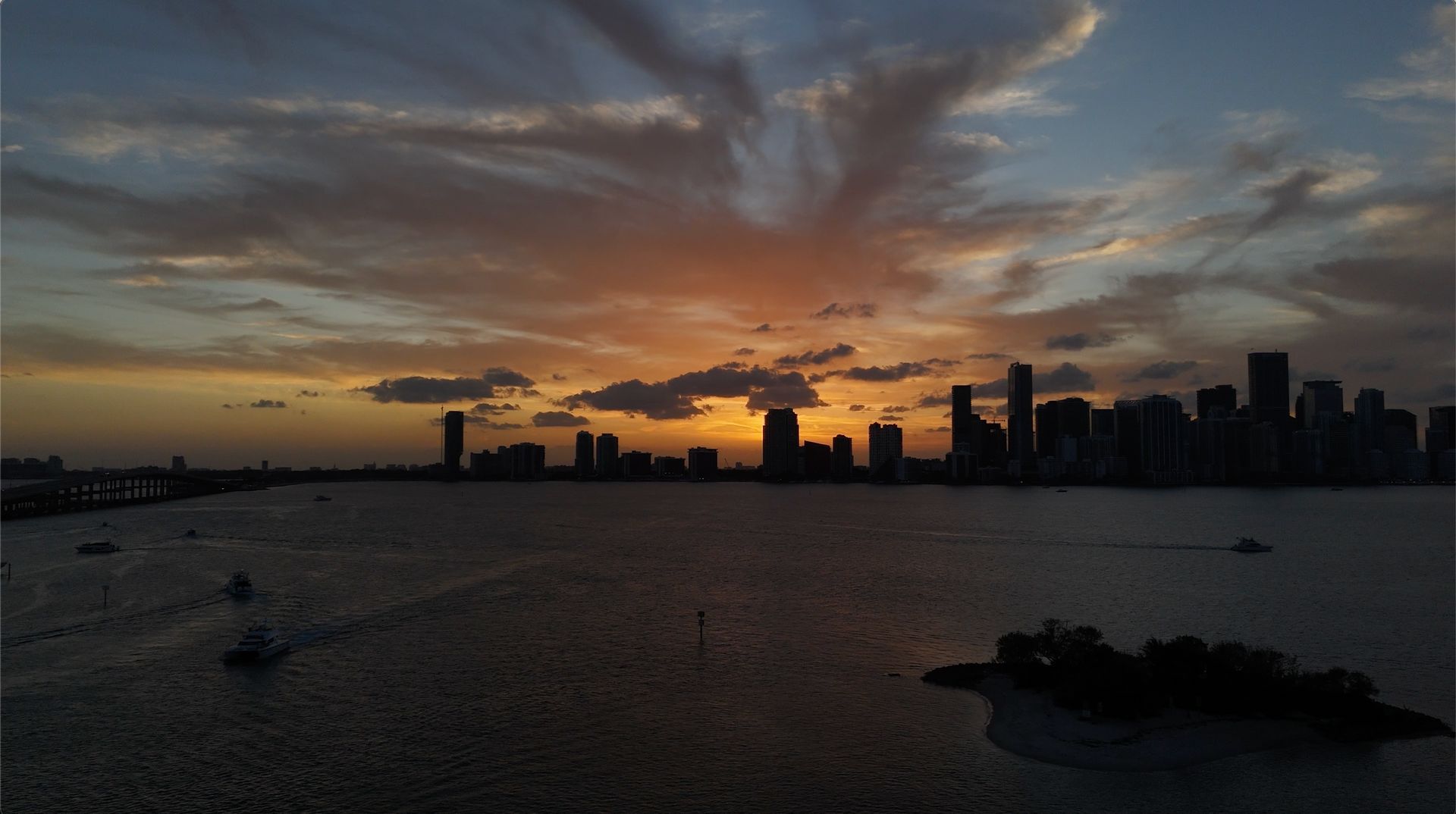 A dramatic Miami sunset skyline captured from above Biscayne Bay with boats cruising across the water during golden hour.