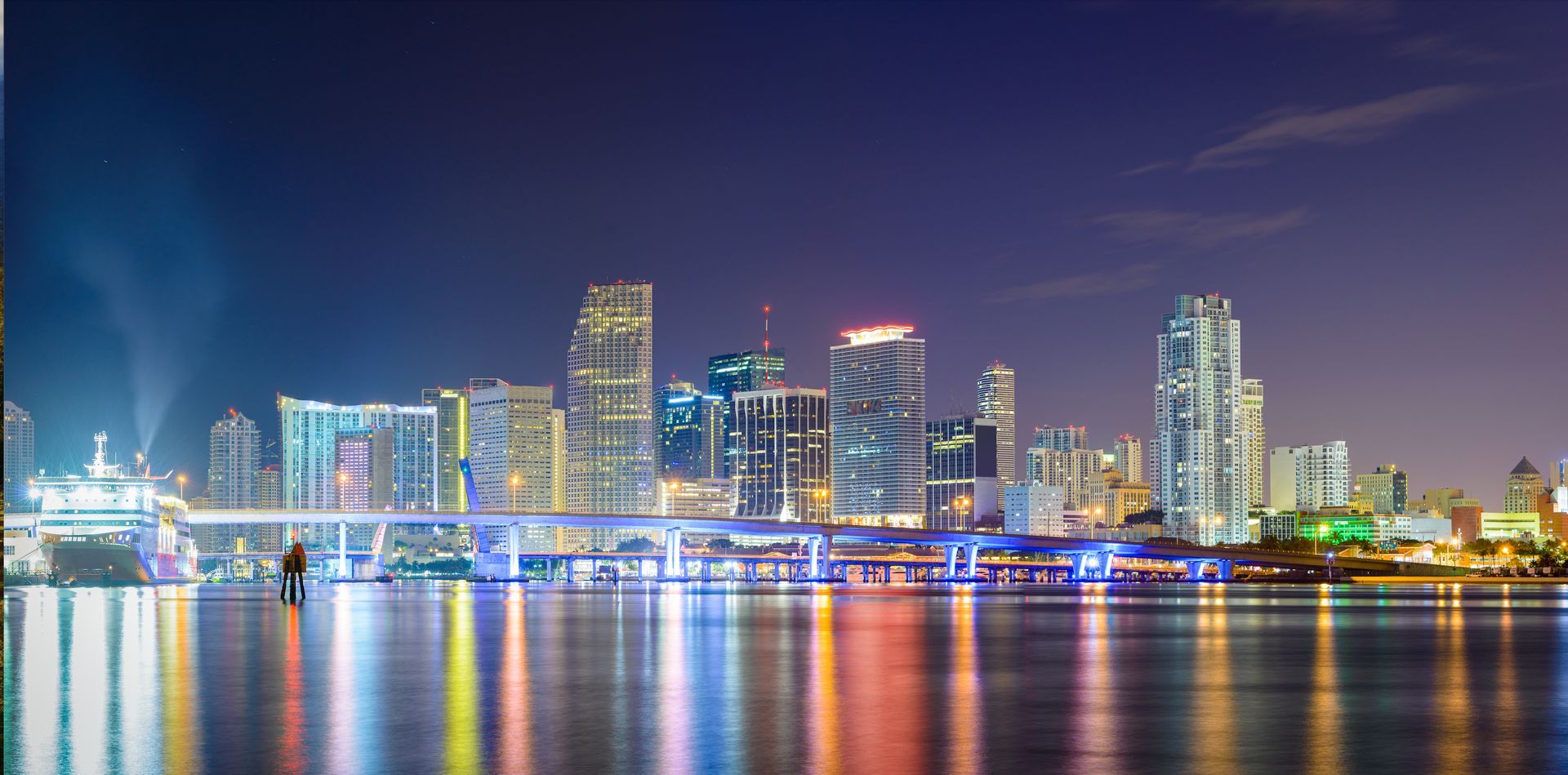 Nighttime Miami skyline with illuminated downtown buildings and colorful reflections on Biscayne Bay.
