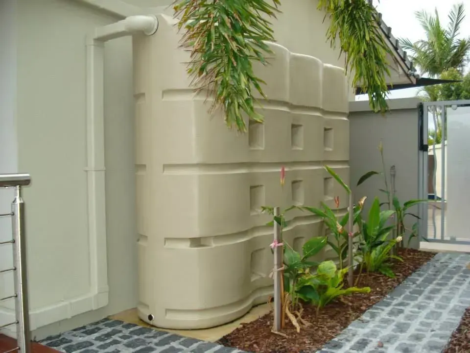 Beige Rectangular Water Tank Next to A White Wall, with A Small Garden in Front — Gladstone Garden & Landscaping Supplies In Burua, QLD