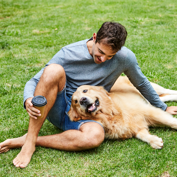 A person sits barefoot on a grassy lawn, petting a golden retriever while holding a coffee cup.