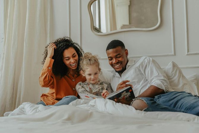 Three people smiling while sitting on a bed reading a book together in a bright, white-walled room.