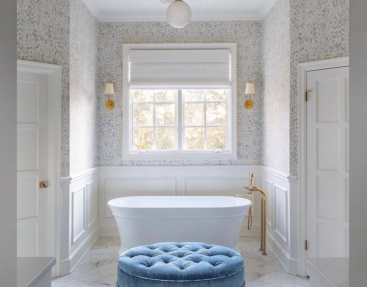 A white freestanding tub sits before a window with blue floral wallpaper and white wainscoting, featuring a blue ottoman.