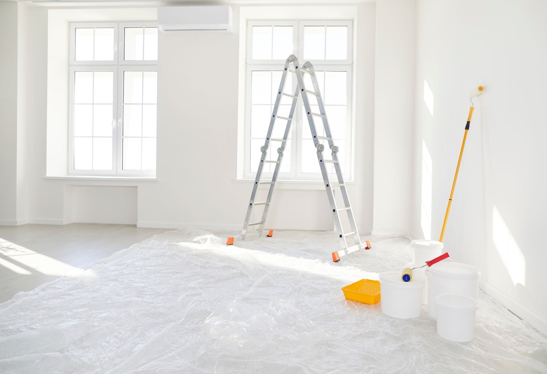 A stepladder, paint buckets, and a roller inside a bright, empty room prepared for painting with a plastic drop cloth.